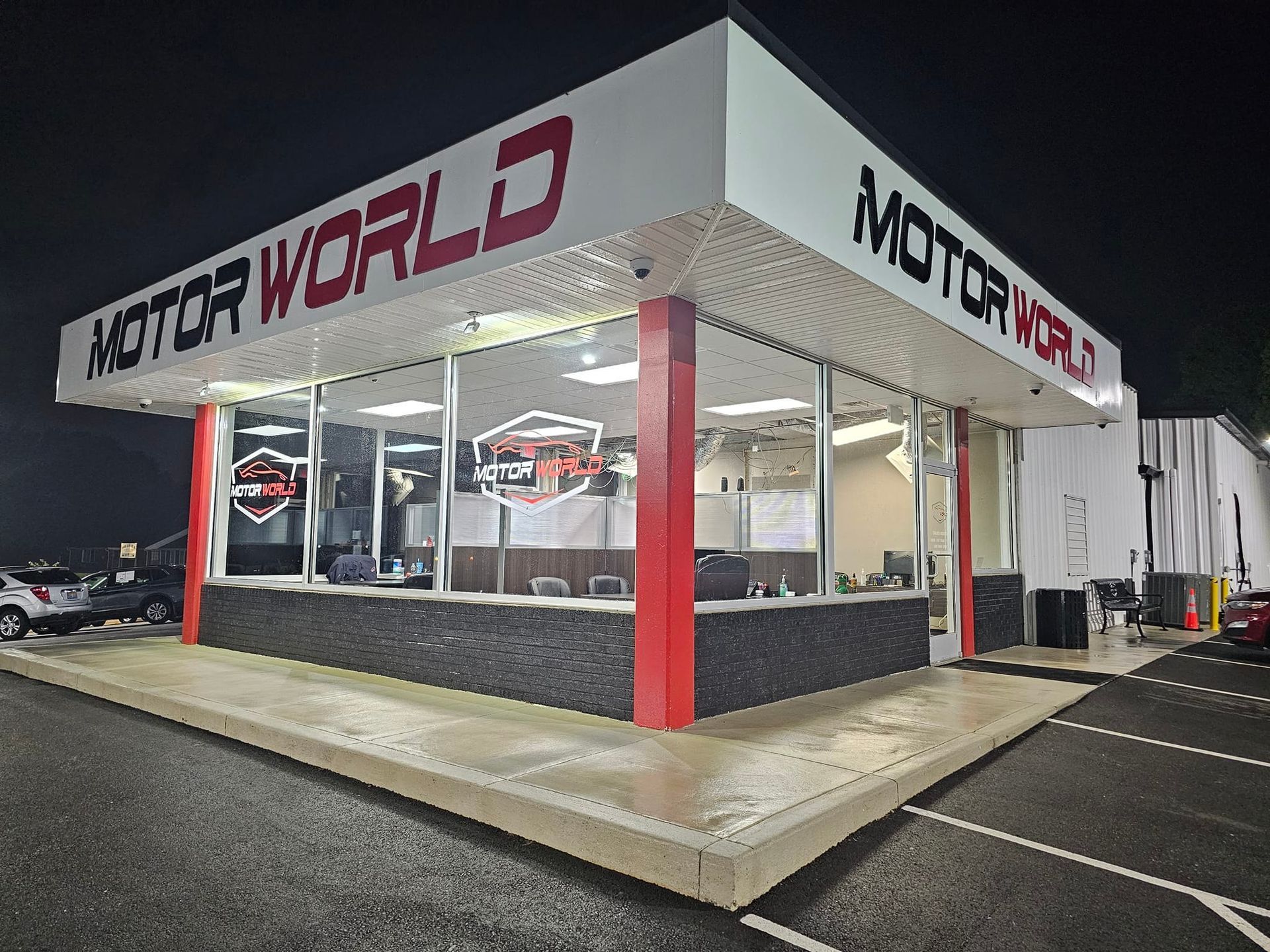 Nighttime exterior of Moto World car dealership with white and red facade and lit showroom windows