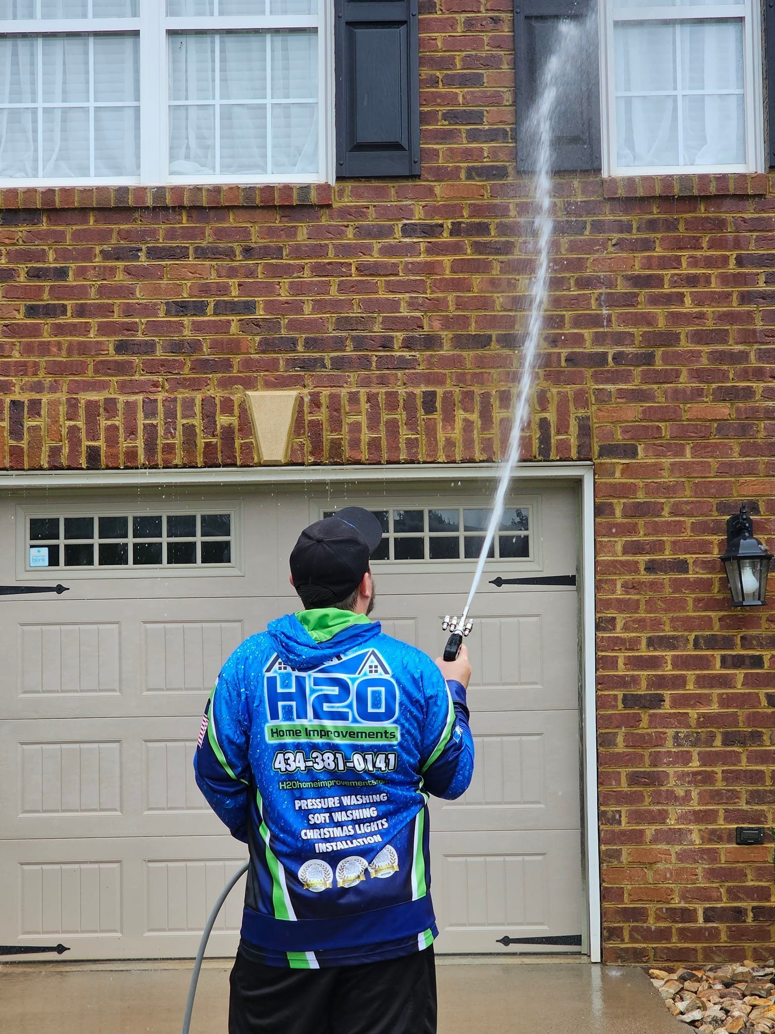 Worker in blue H2O jacket pressure-washing a brick house near a garage door