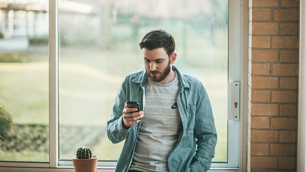 Man in a denim shirt looking at his phone by a window with a small potted plant