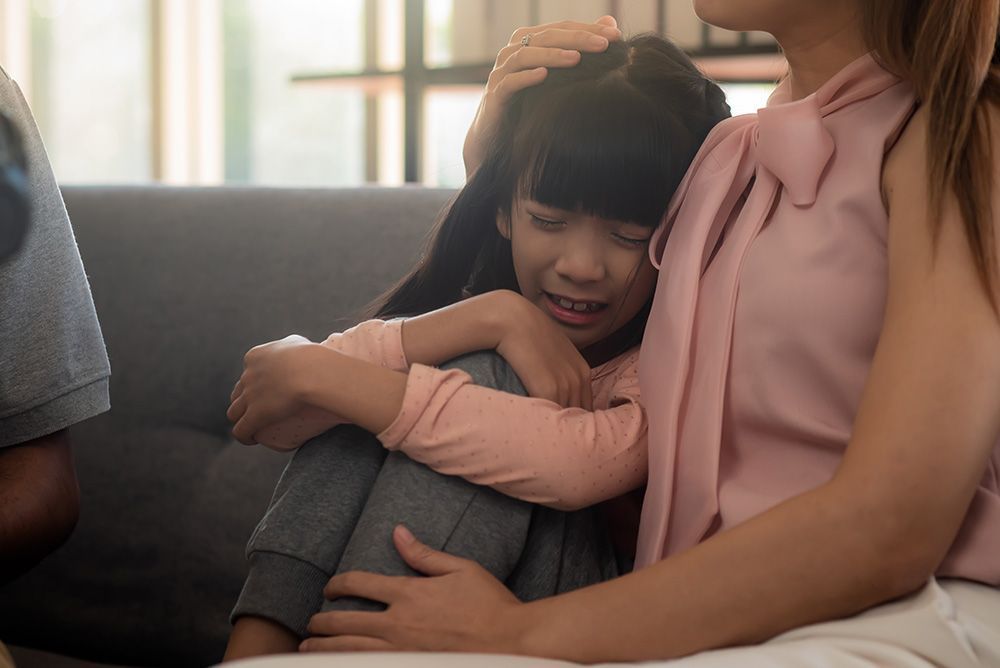 Child crying while seated on an adult's lap, comforted by another adult on a couch.