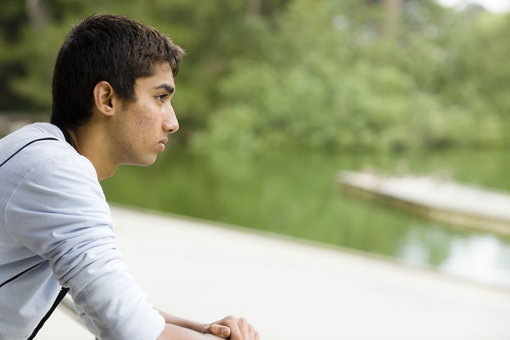 Person in a light shirt leaning on a railing beside a calm pond, looking thoughtfully ahead.
