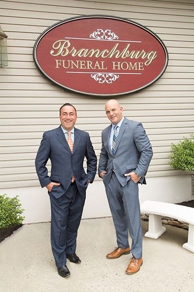 Two men in suits stand outside Branchburg Funeral Home beneath a red oval sign.