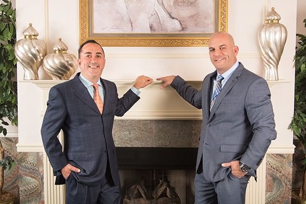 Two men in suits posing beside a fireplace with decorative gold vases and a framed painting.