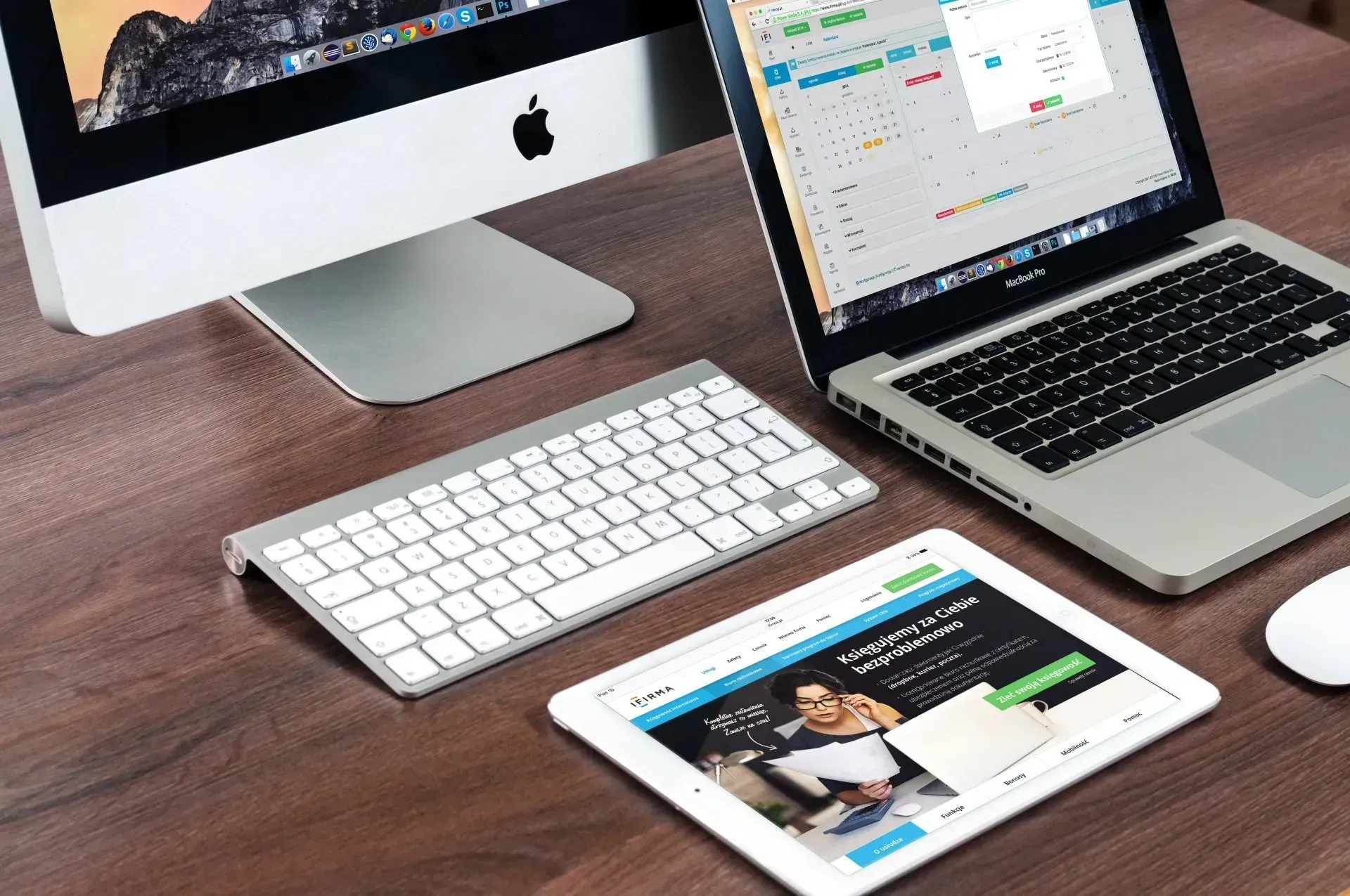 Desktop workspace with iMac, laptop, keyboard, and tablet on a wooden desk