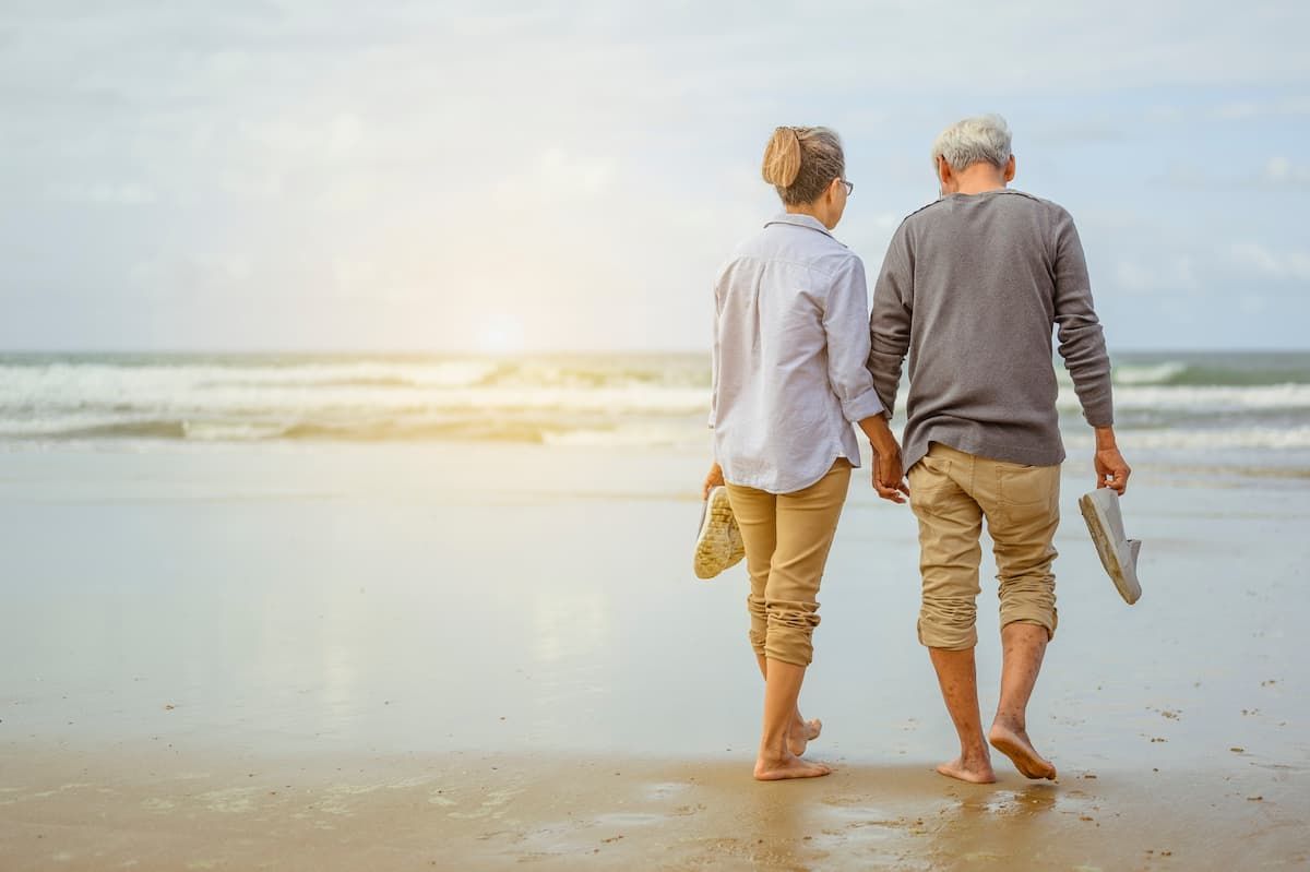 Two people walking barefoot on a beach, holding shoes, with the ocean in the distance