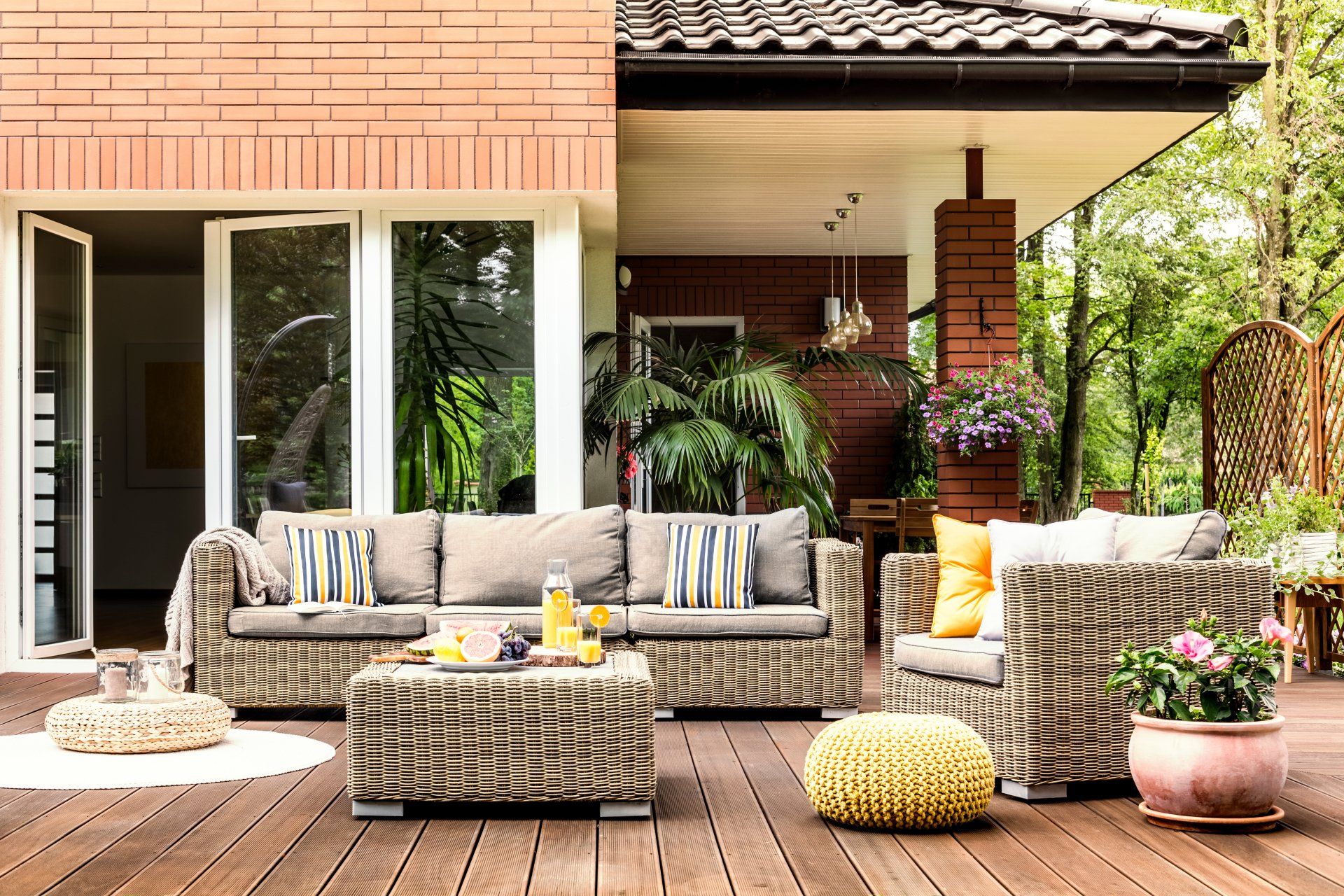 Yellow pouf next to a rattan armchair and flowers on wooden patio with striped pillows on sofa