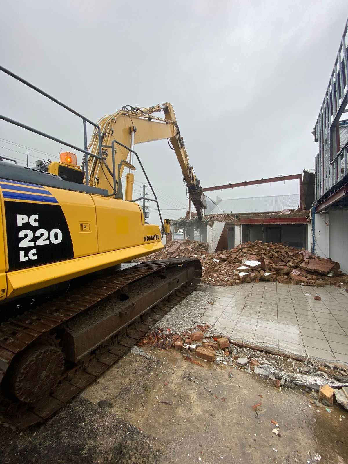 Excavator demolishing building on site with rubble piles and exposed steel framing.