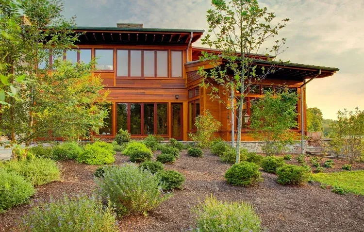 A large wooden house with a lot of windows is surrounded by trees and bushes.