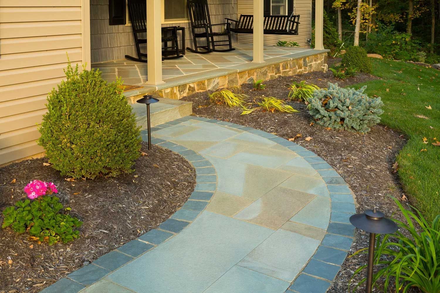 A stone walkway leading to a porch with rocking chairs.
