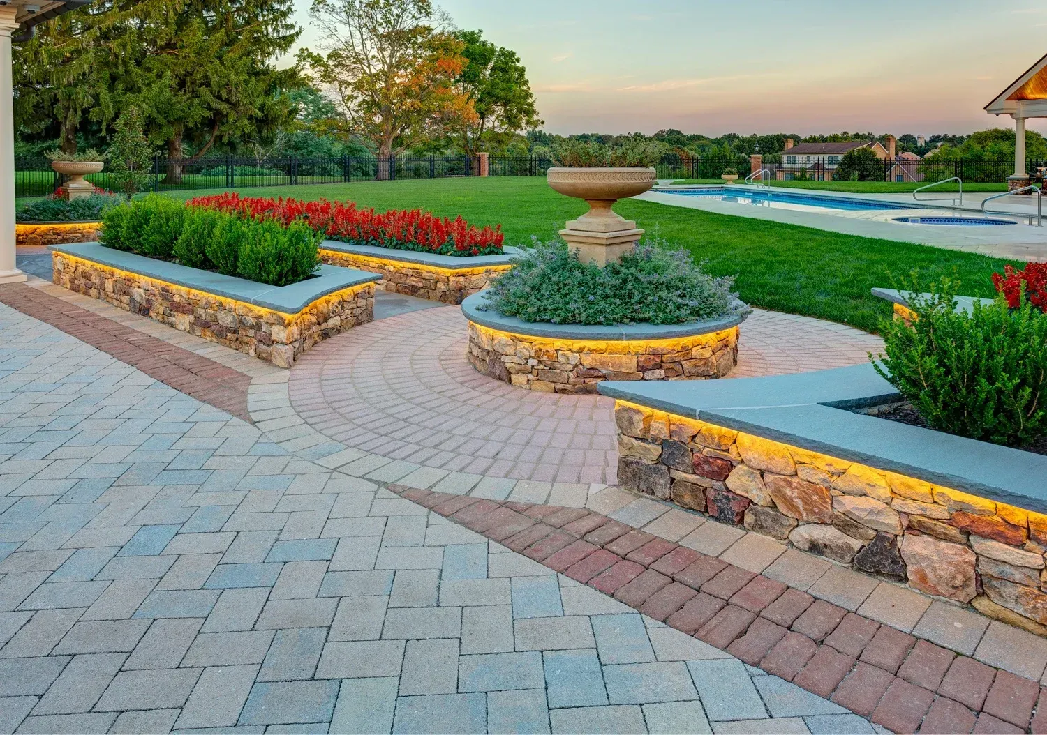 A brick walkway with a fountain in the middle and a swimming pool in the background.