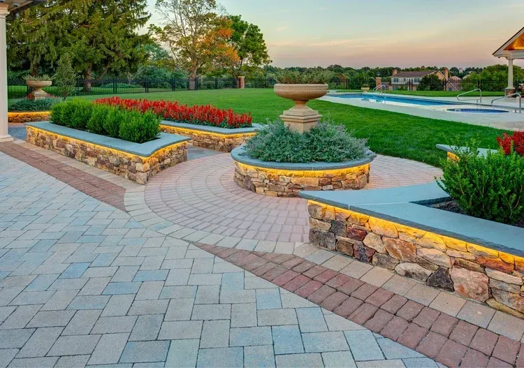 A brick walkway with a fountain in the middle and a swimming pool in the background.