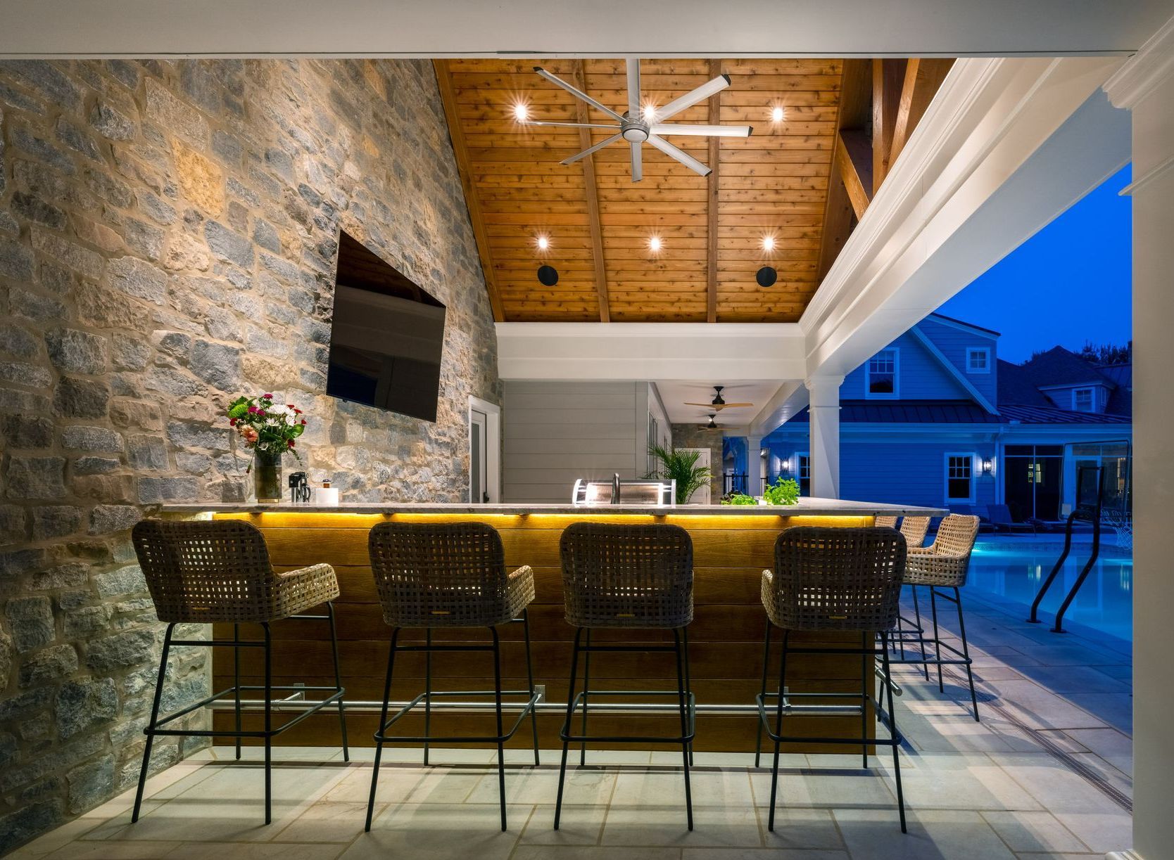 Outdoor bar with stone wall, wooden ceiling, bar stools, and pool in the background.