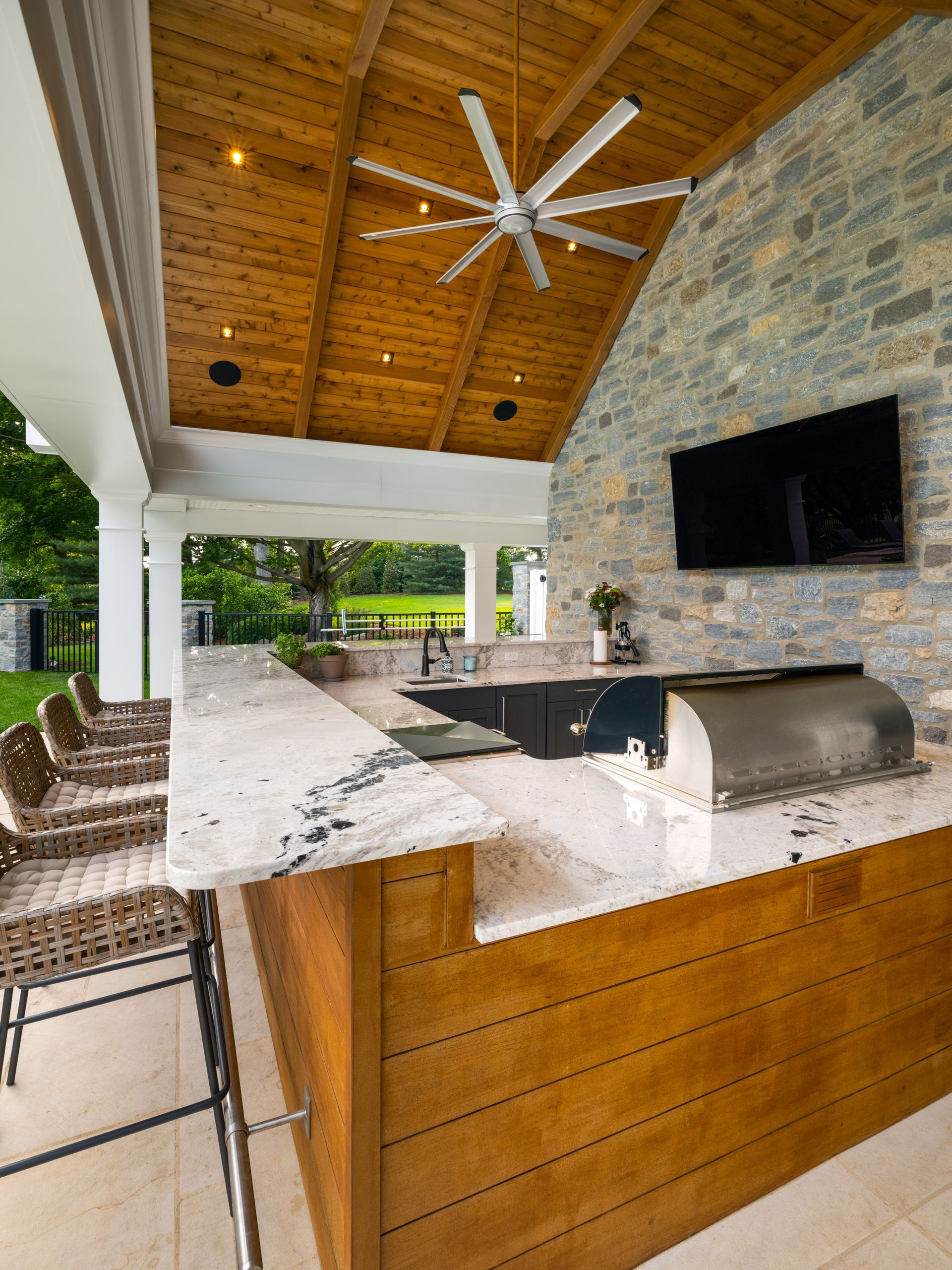 Outdoor kitchen with stone wall, wood ceiling, grill, granite countertops, and seating.