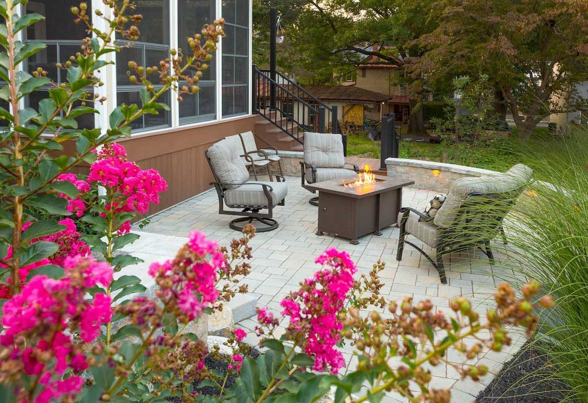 A patio with chairs , a table and a fire pit surrounded by pink flowers.