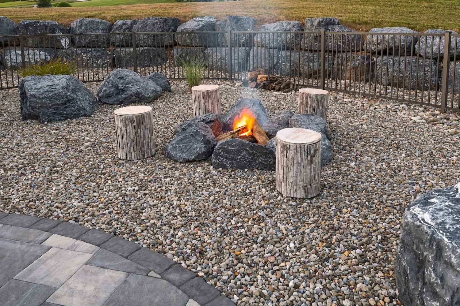 A fire pit is surrounded by rocks and stump stools in a gravel area.