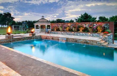 Swimming pool with a gazebo, brick wall with flowers, and evening sky.