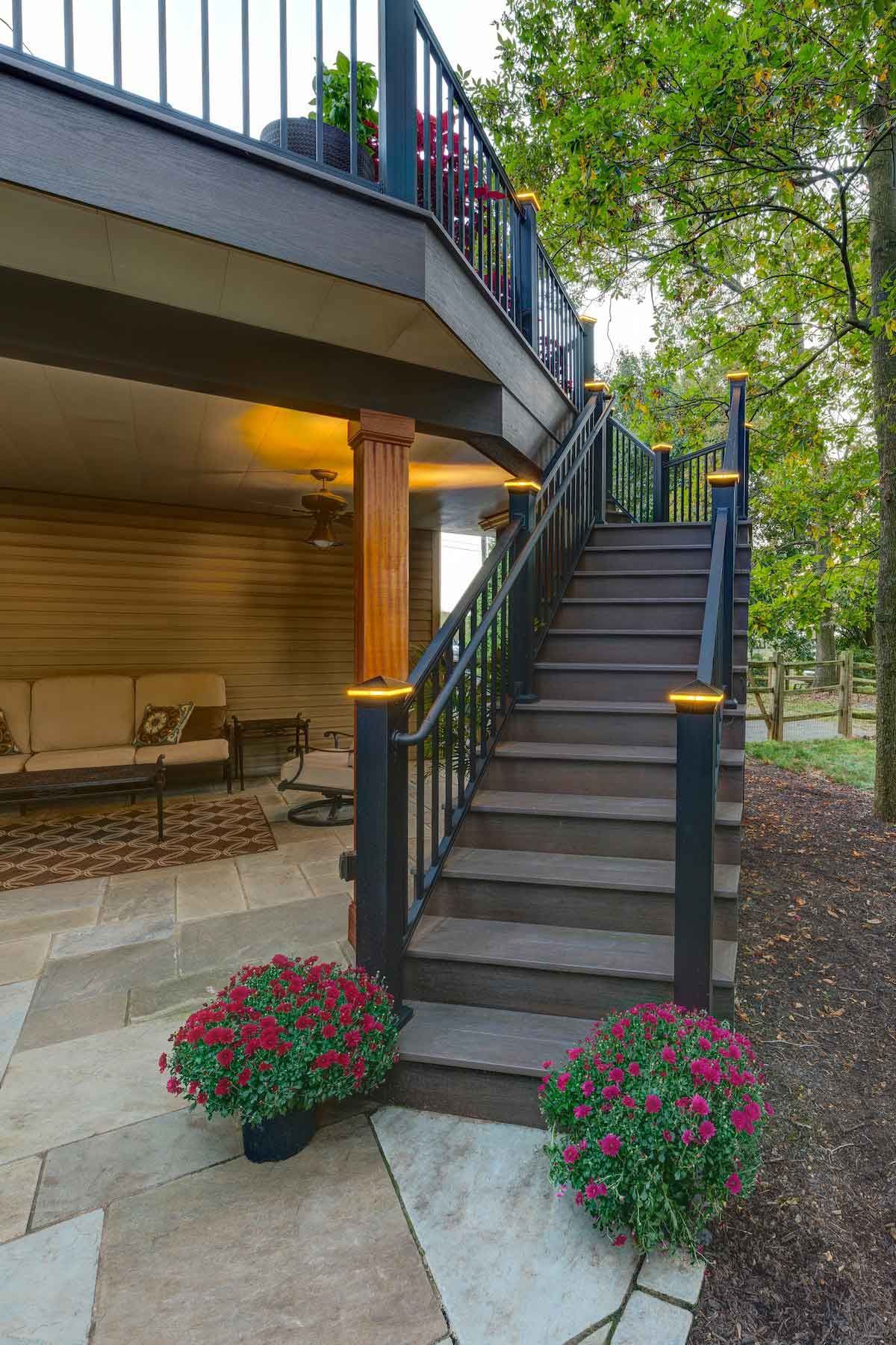 A deck with stairs leading up to it and flowers on the sidewalk.