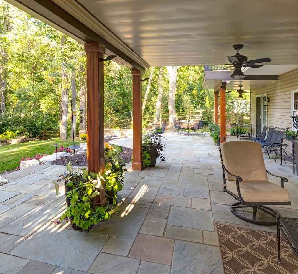 Covered patio with stone floor, seating, and potted plants overlooking a yard with trees.