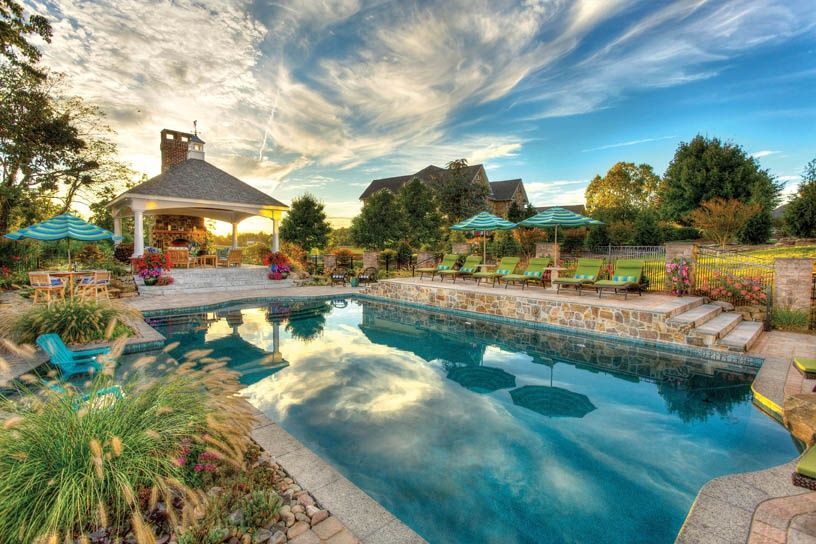 Sunny backyard pool with stone patio, lounge chairs, gazebo, and reflected clouds at sunset