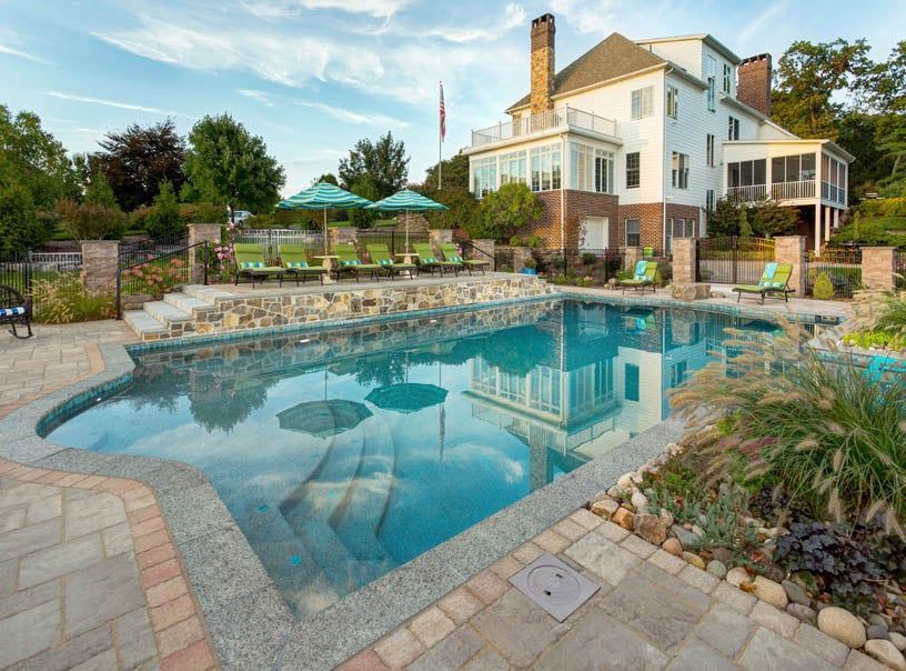 A large swimming pool is surrounded by chairs and umbrellas in front of a large house.