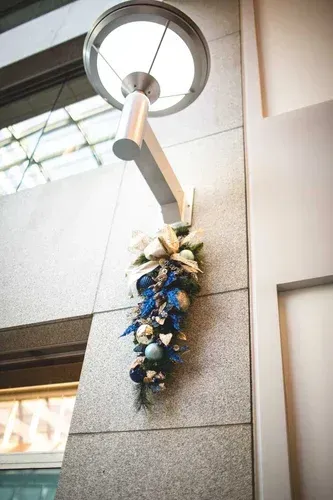 Festive holiday decoration hangs below a modern lamp on a gray wall, with blue and silver ornaments.