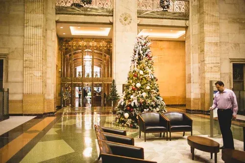 Grand lobby with Christmas tree. Man near chair, ornate gold doors. Marble columns, patterned floor.