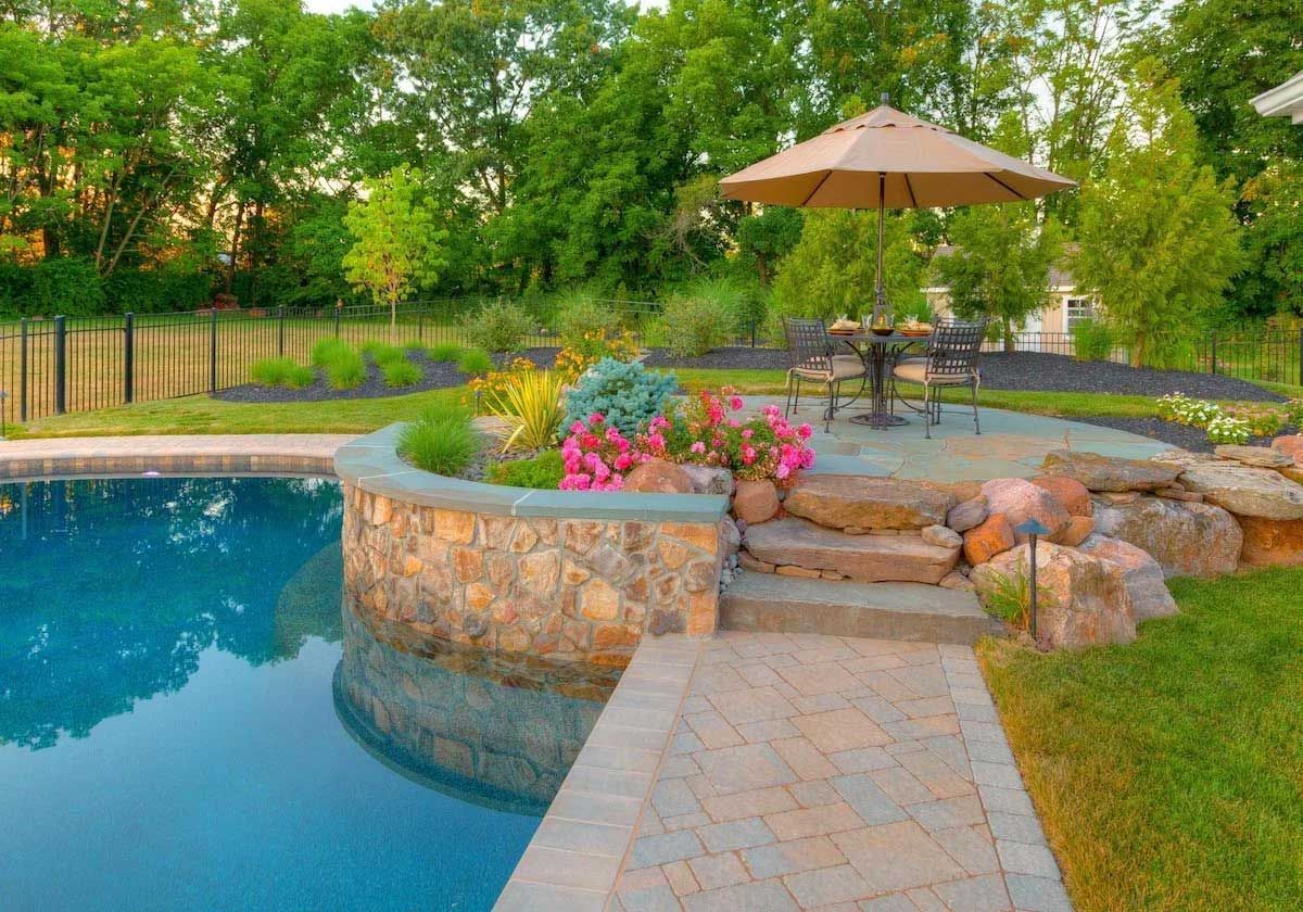 There is a swimming pool in the backyard with a table and chairs and an umbrella.