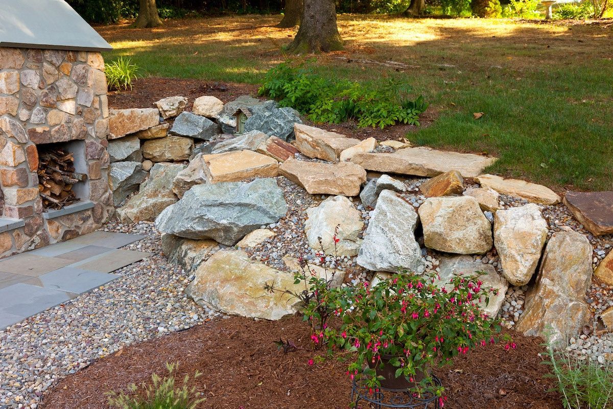 A stone fireplace is surrounded by rocks and flowers in a backyard.