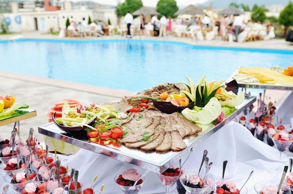 A buffet table with food and drinks in front of a swimming pool.