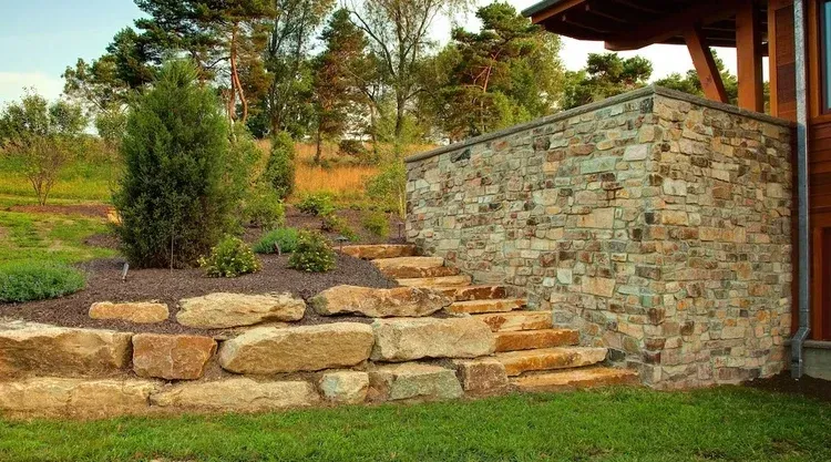 A stone wall with stairs leading up to it is in the backyard of a house.