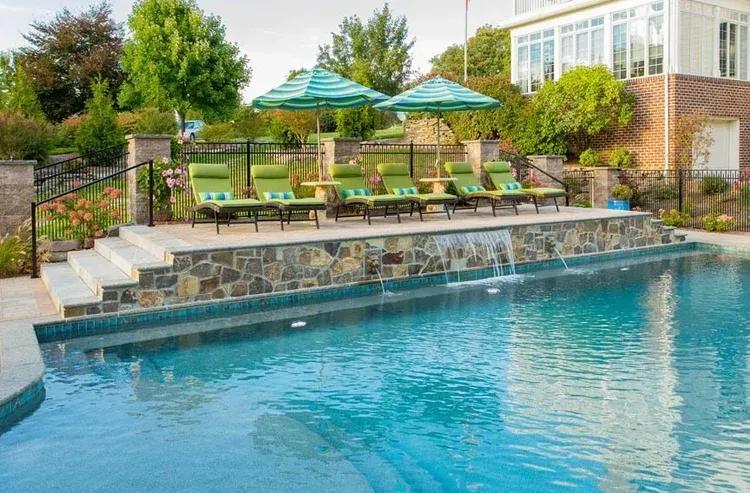 A large swimming pool with green chairs and umbrellas in front of a house.
