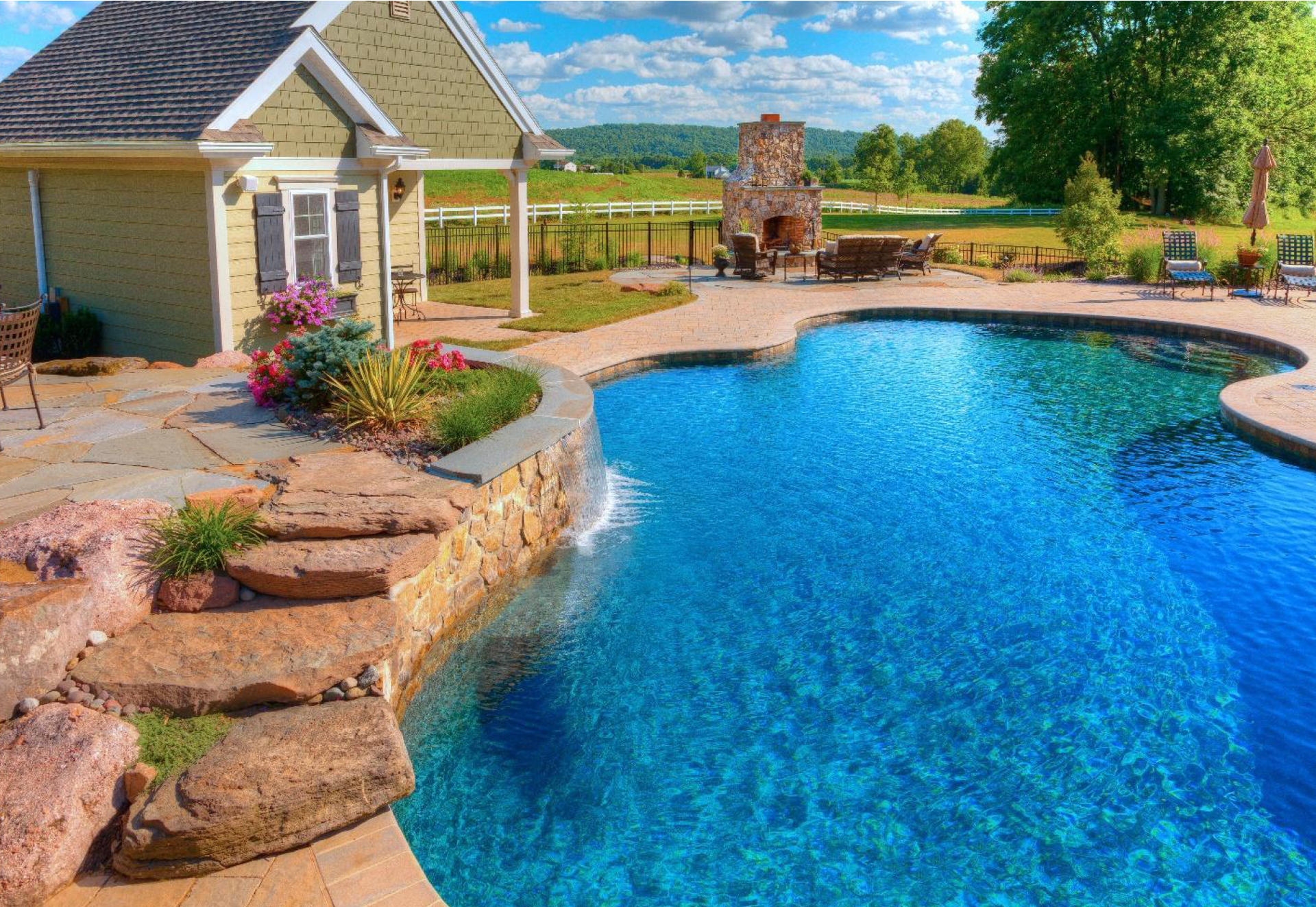 Backyard swimming pool with stone deck, flower beds, and patio beside a house under a bright blue sky