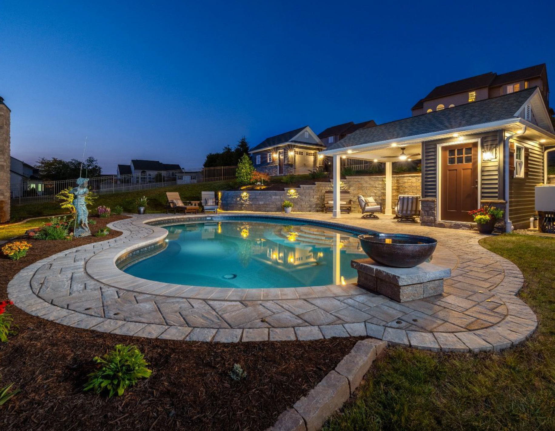 Backyard patio with lit swimming pool and stone deck beside a house at dusk