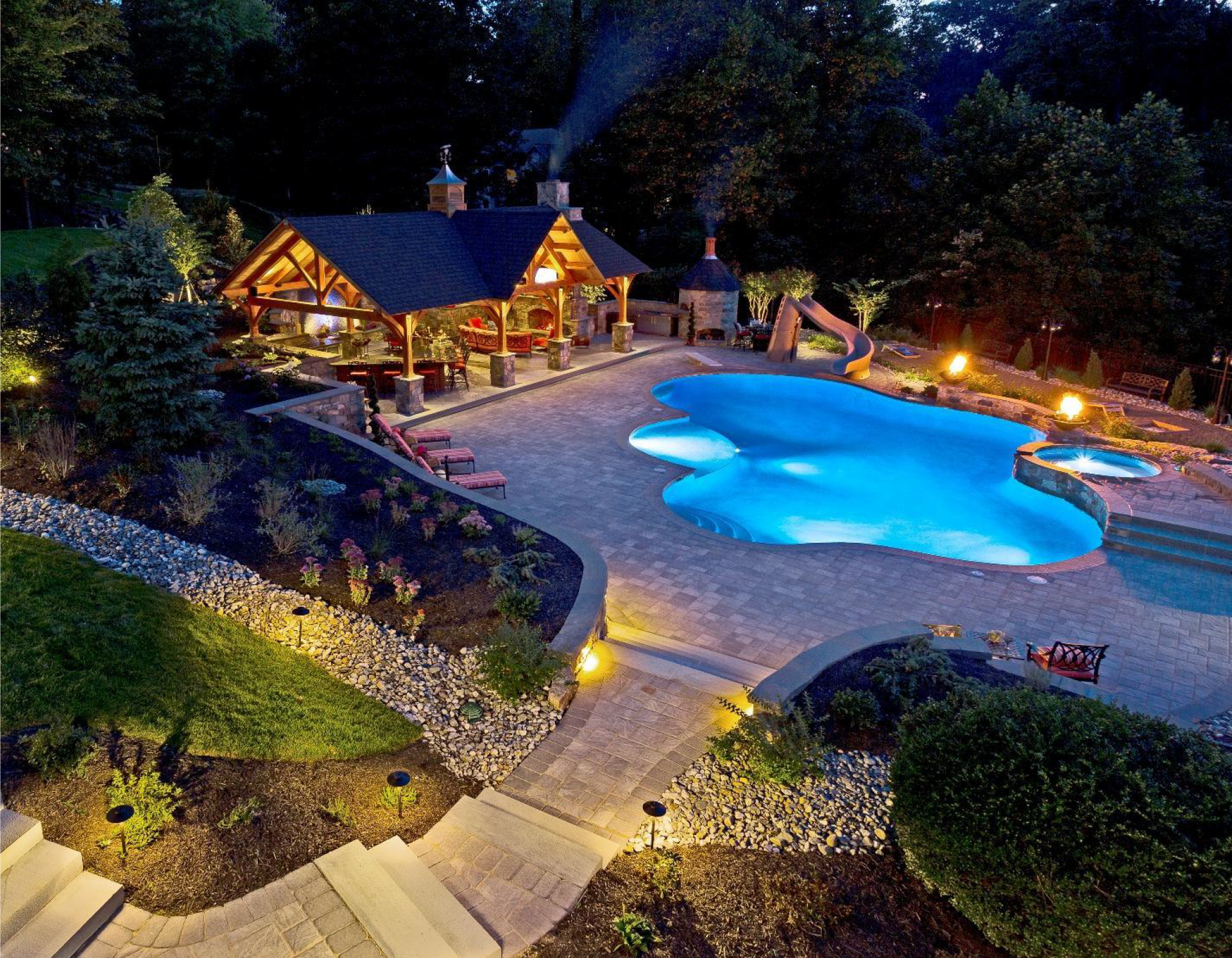 Nighttime view of a lit lodge beside a glowing blue swimming pool in a wooded backyard