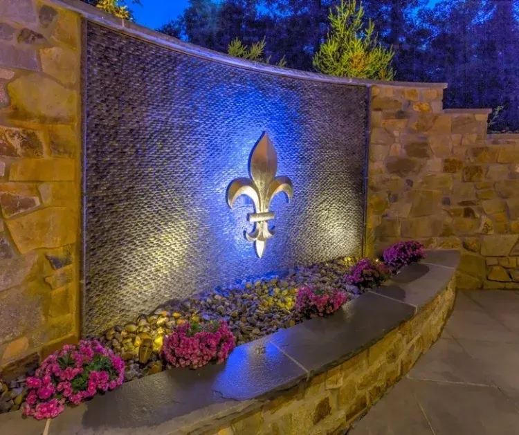 A water fountain with a fleur de lis on it is surrounded by rocks and flowers.