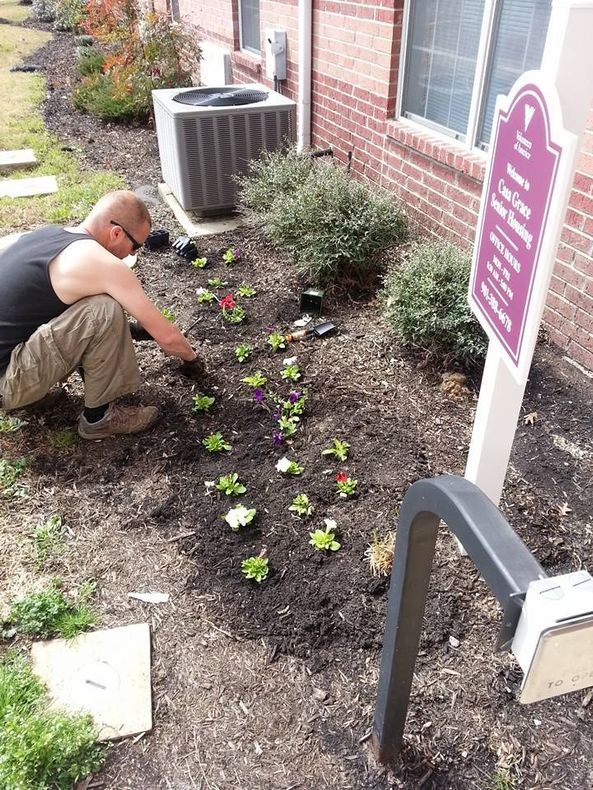 Person planting flowers in a garden bed next to a brick building.