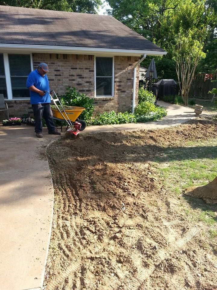 Person using a tiller to cultivate soil near a house. Pathway on the left and landscaping on the right.