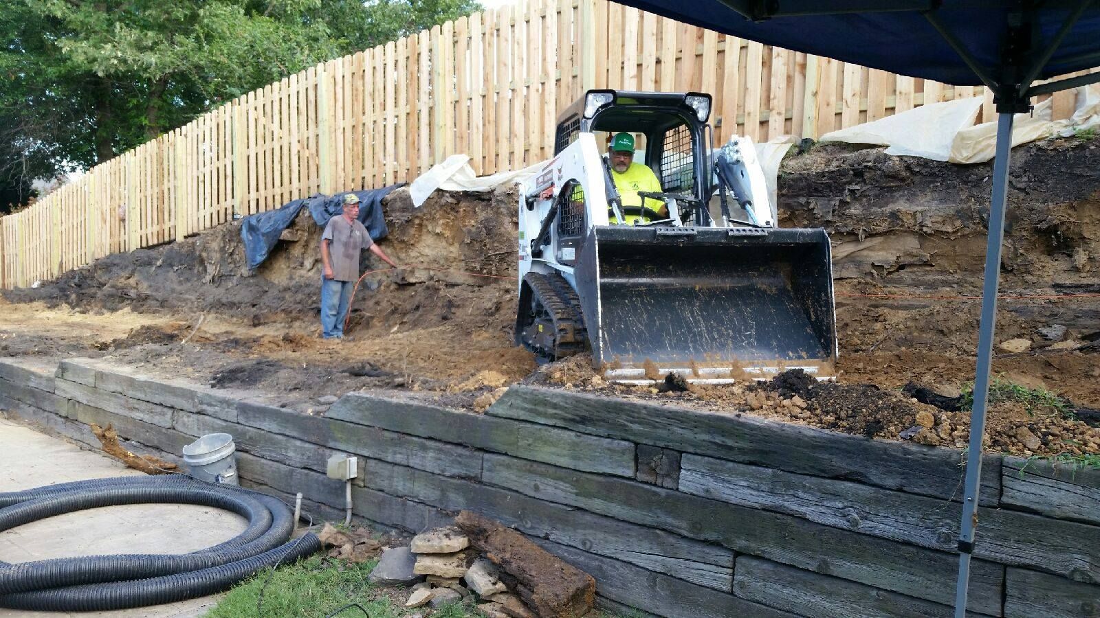 Construction site with a skid steer and a worker near a wooden retaining wall and fence.