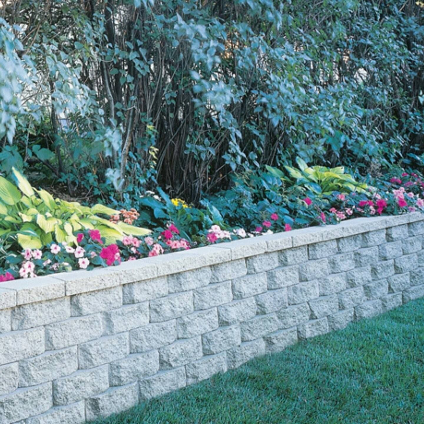 Retaining wall with a bed of flowers and plants; lawn in front, trees in the background.