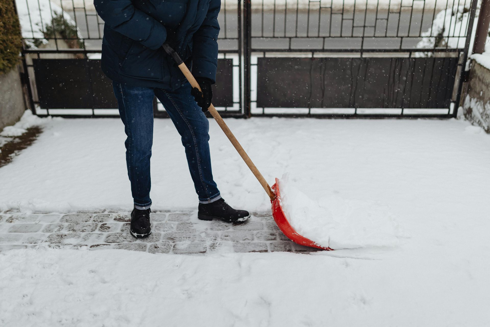 Person shoveling snow from a brick pathway in front of a metal gate.