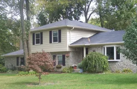 Two-story suburban house with beige siding, brick accents, and trees in the front yard