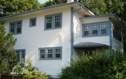 White two-story house with gray trim, multiple windows, and a front porch surrounded by greenery