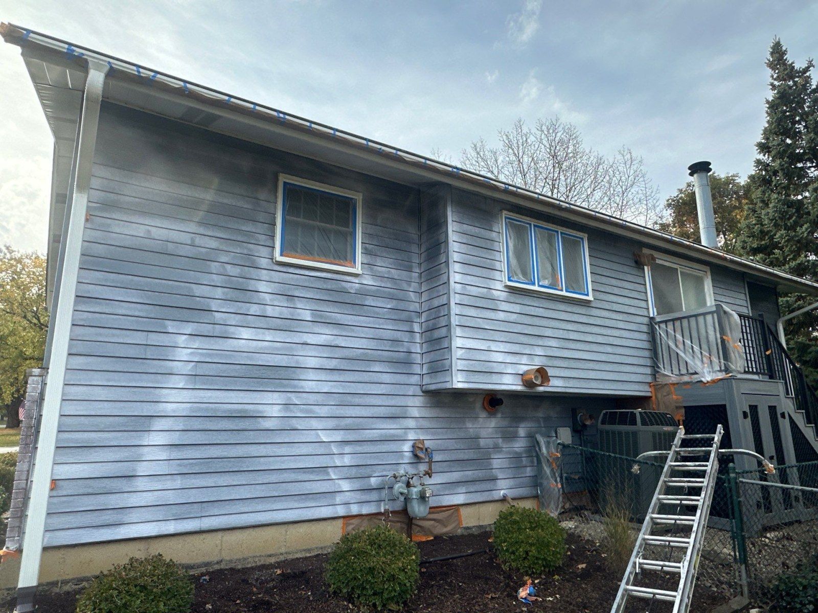 Blue-gray two-story house with peeling siding and an extension ladder leaning against the deck entrance