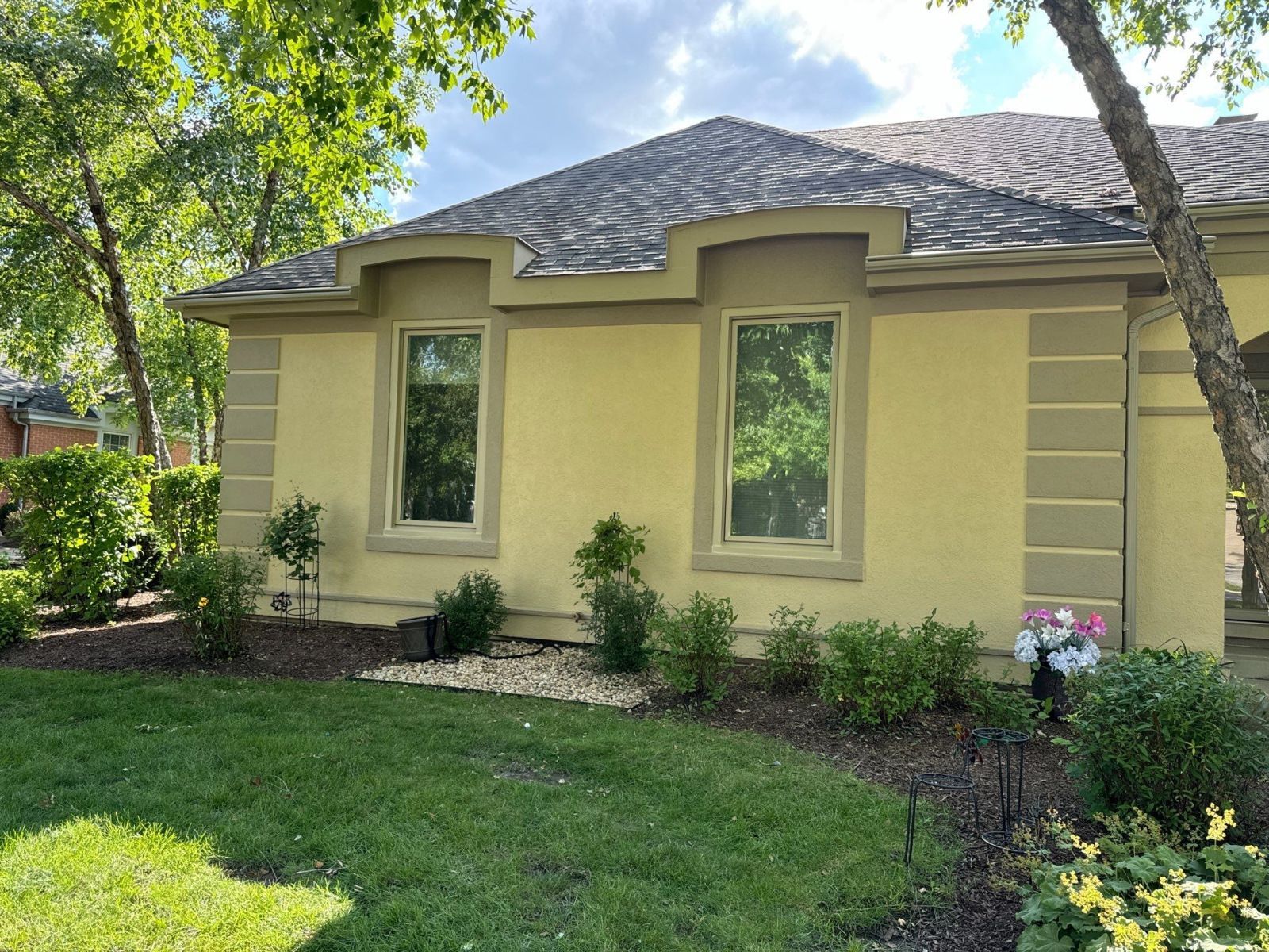 Single-story beige house with a shingled roof, front windows, and a green lawn under leafy trees