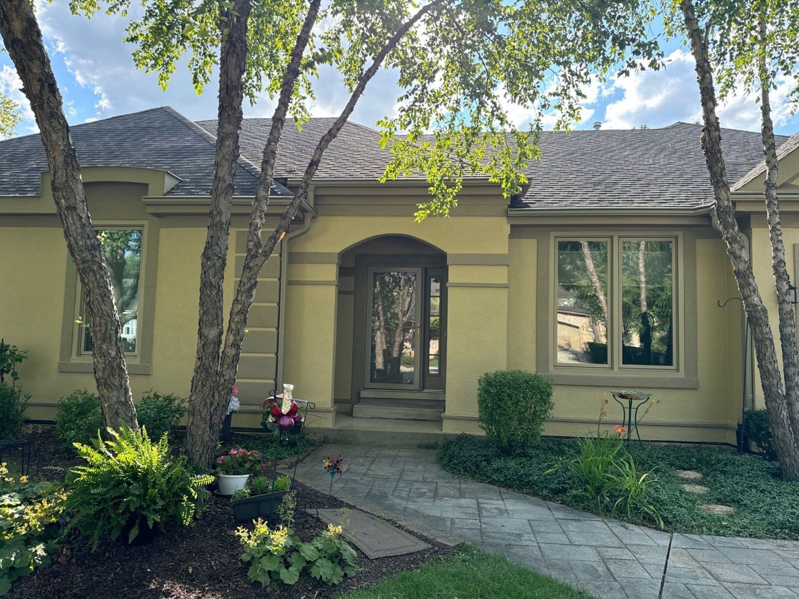 Front entrance of a beige house with arched doorway, trees, shrubs, and a stone walkway in front.