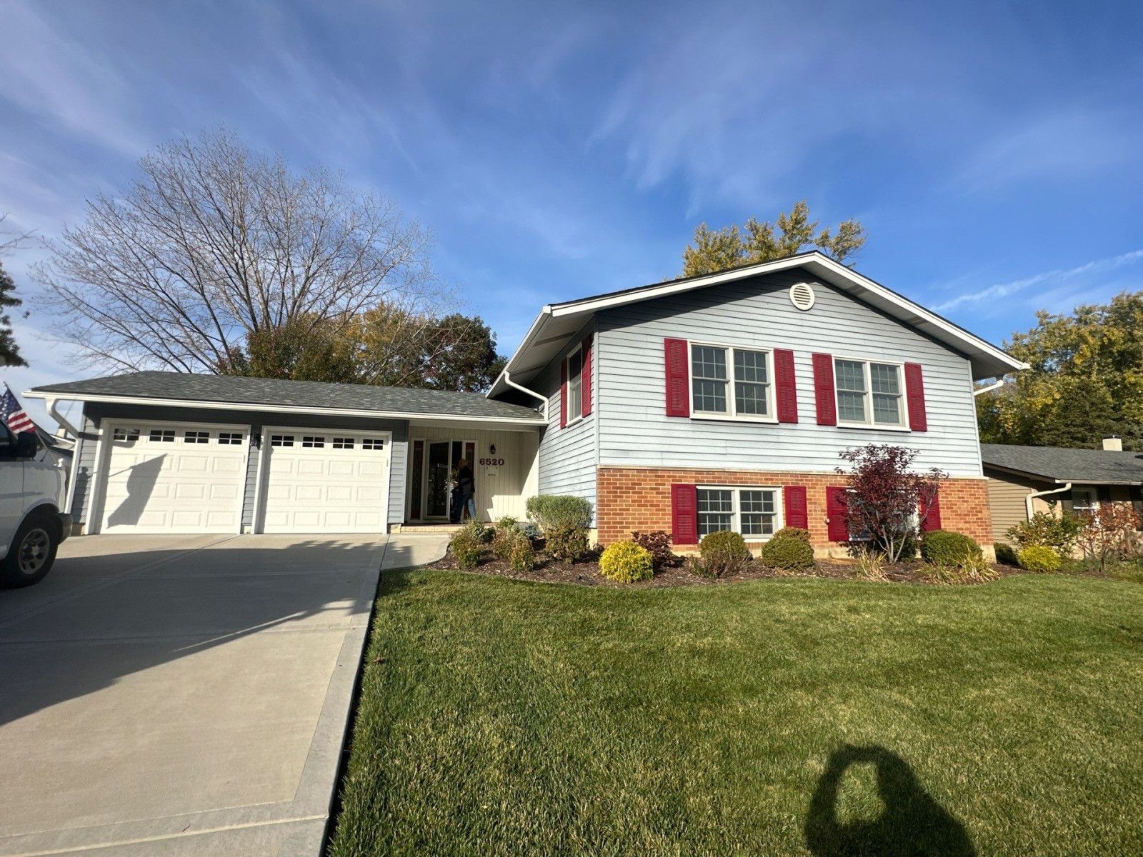 Suburban house with white siding, red shutters, attached garage, and a concrete driveway under a blue sky