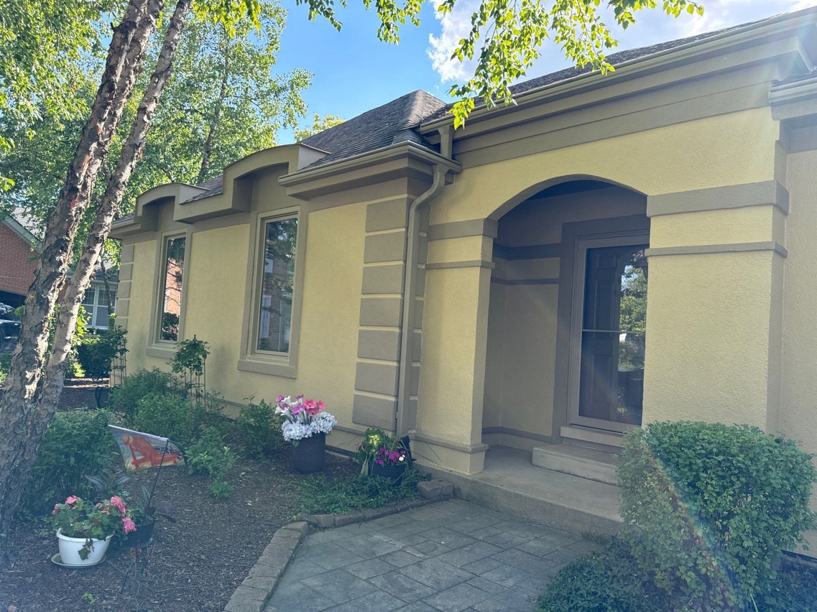 Stucco house exterior with arched entry, windows, and flower pots in a shaded front yard.