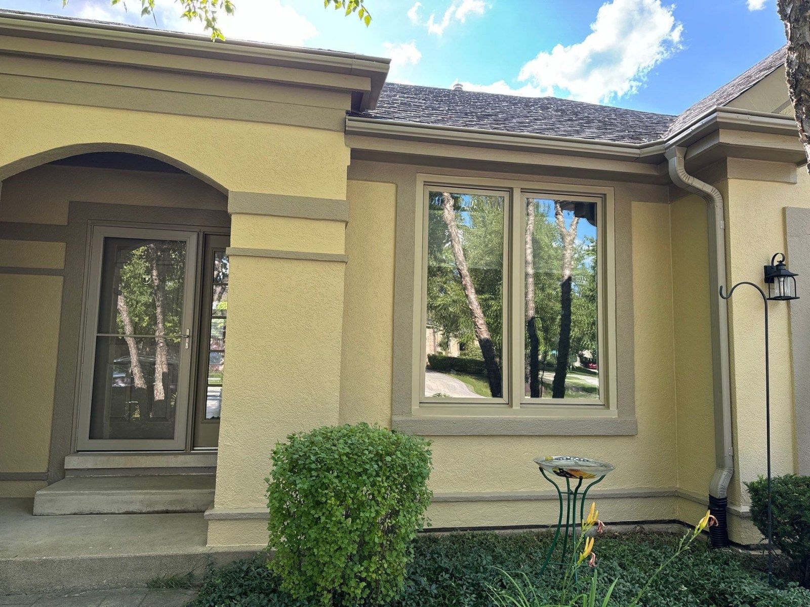 Front exterior of a tan stucco house with windows, a front entry, and a small shrub by the wall