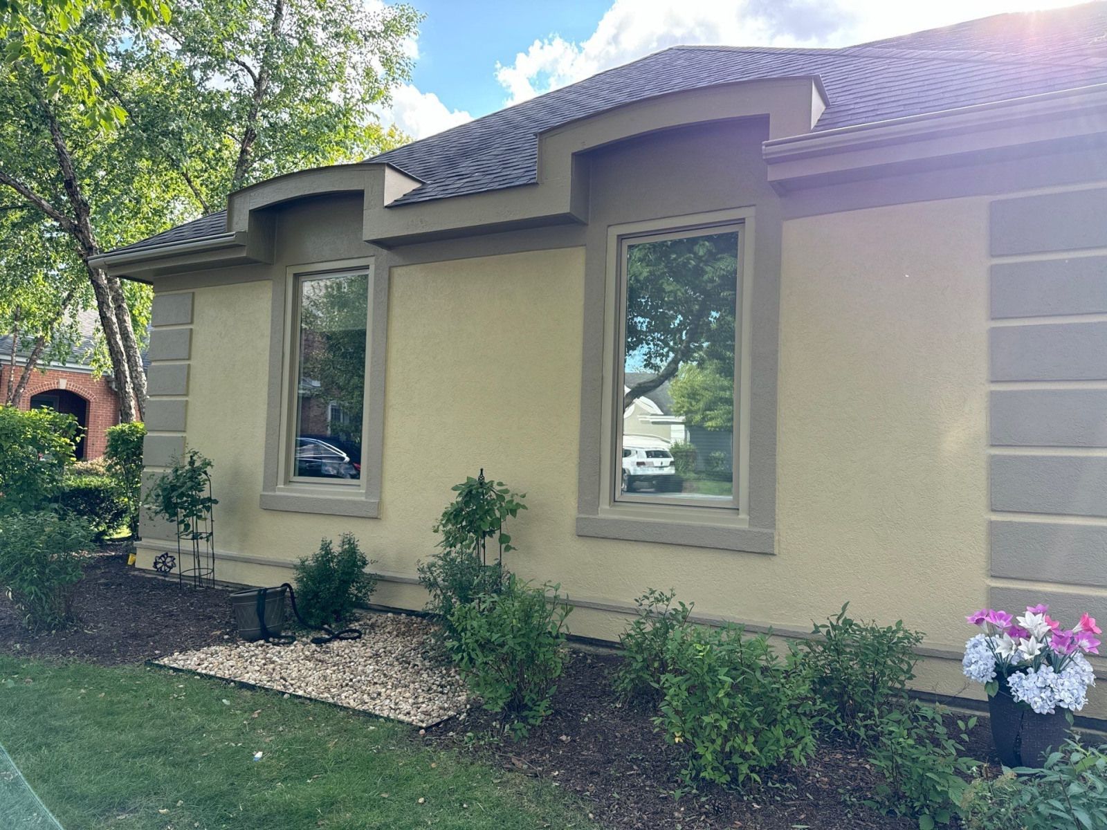Single-story tan house with green trim, front windows, shrubs, and a small flower pot by the entry