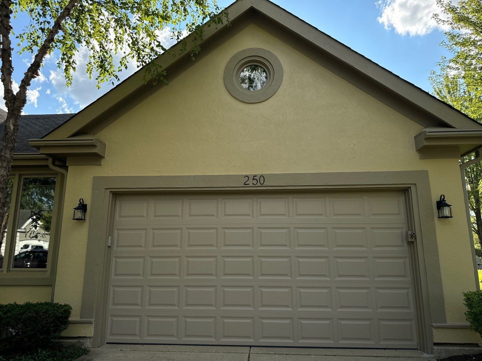 Front view of a beige garage with a closed door, circular attic window, and trees framing the house